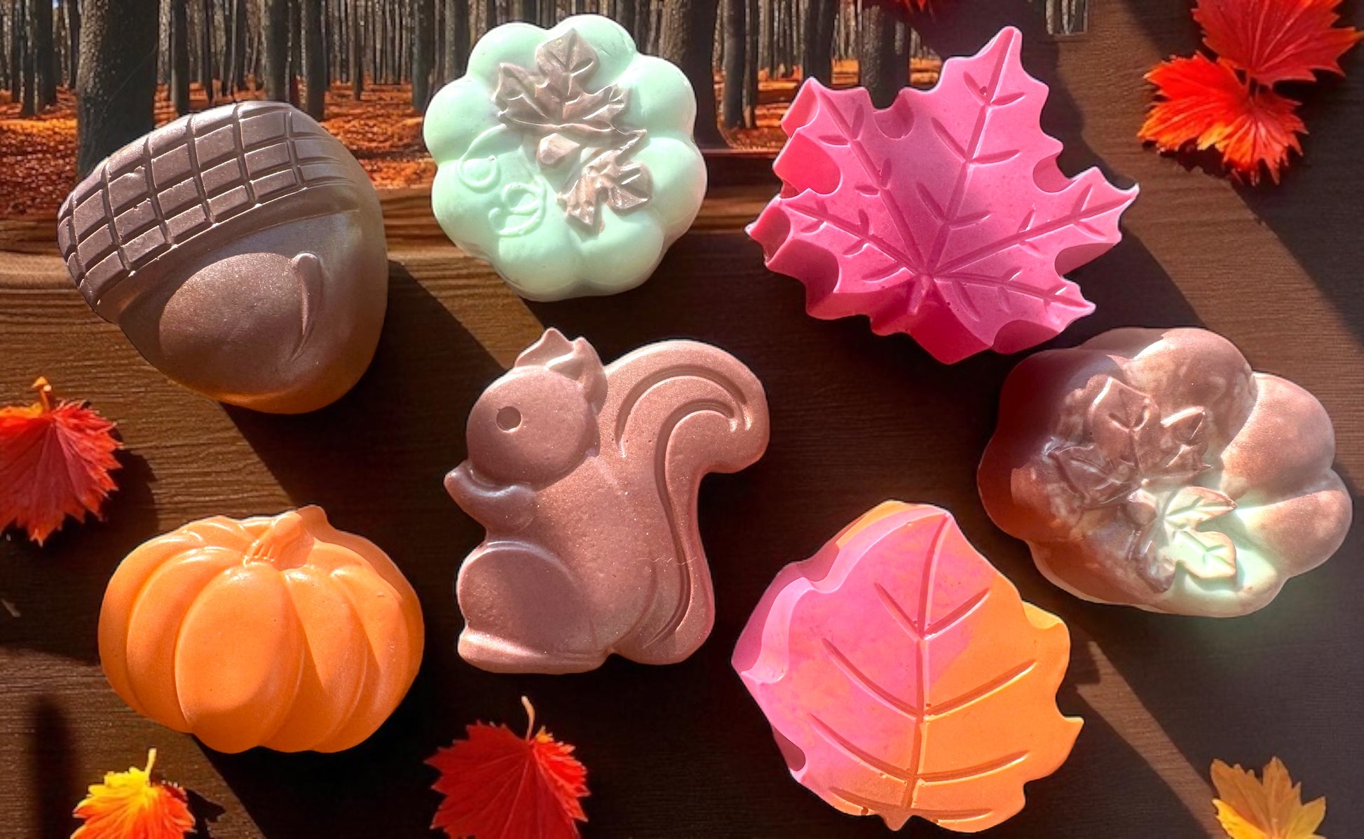 Colorful soap bars shaped like pumpkins, leaves, and a squirrel on a wooden surface with autumn leaves.