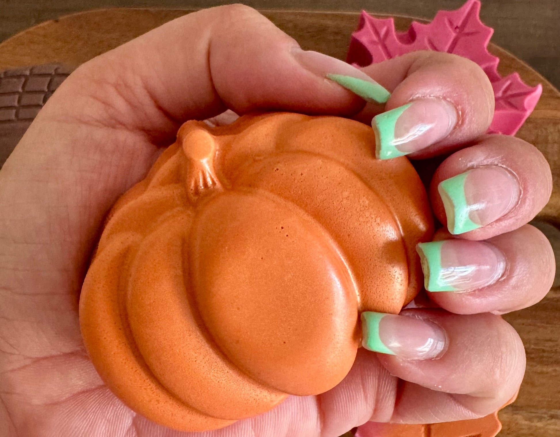 Hand holding a small orange pumpkin-shaped object with pink leaf-shaped objects in the background.