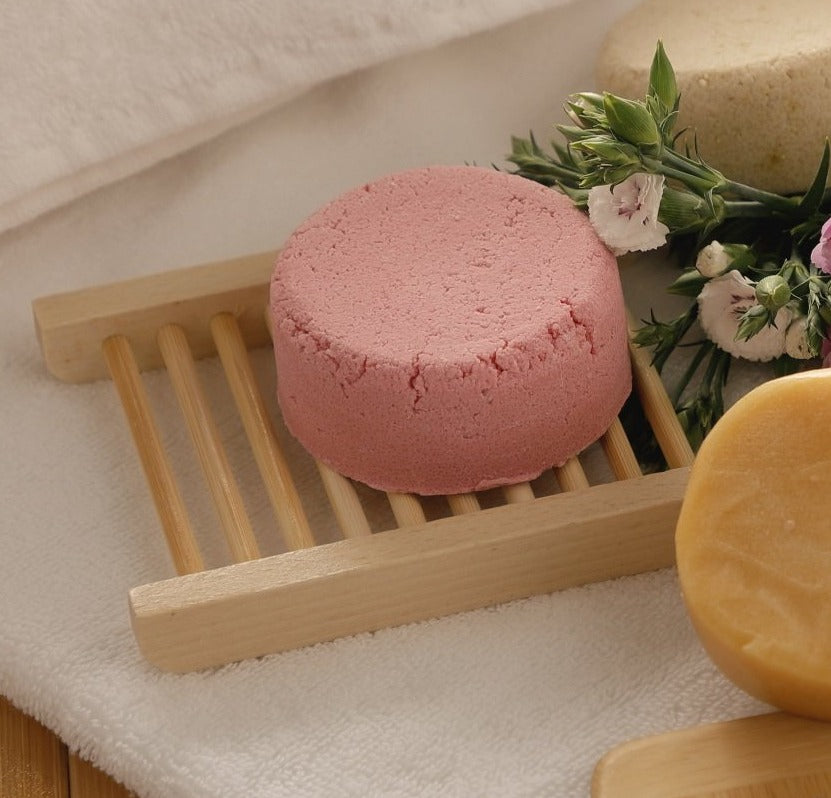 Pink bath bomb on a wooden soap dish with flowers and another yellow bath bomb in the background.