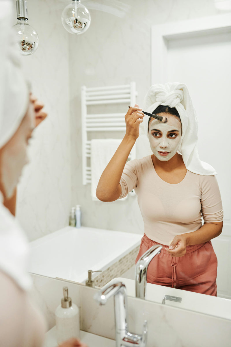 Woman applying facial mask in front of a mirror in a bathroom.